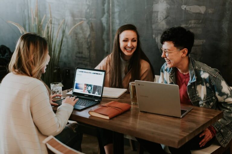 About Us Sell My Website About Us Image of two girls and on guy laughing while on laptops at a table in a bar.